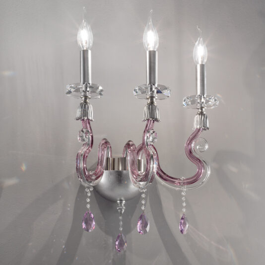 The Contemporary 3 Arm Candle Style Glass Wall Light features three candle-shaped lights, each with a silver base and clear, ornate glass supports. The sconce's arms are crafted from pink glass adorned with hanging crystal pendants, all elegantly mounted on a silver backing plate.