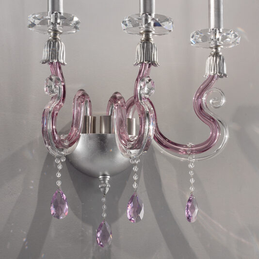 The Contemporary 3 Arm Candle Style Glass Wall Light features elegant, curved arms with pink glass and silver accents, adorned with hanging crystal prisms that catch the light. The base is silver, adding a touch of sophistication to the design.