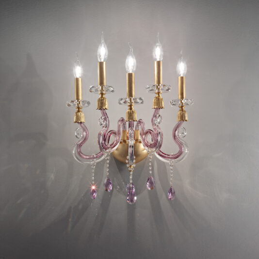 The Contemporary 5 Arm Candle Style Glass Wall Light is a wall-mounted chandelier that boasts gold accents and pink decorative glass arms. It features five candle-shaped lights that illuminate the space, complemented by pink dangling crystal ornaments, which enhance its elegance against a plain, light gray wall.