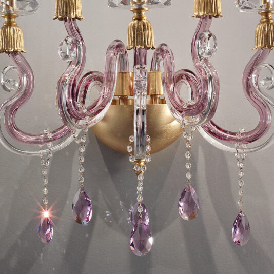 A close-up of the Contemporary 5 Arm Candle Style Glass Wall Light showcases its elegant, modern design with gracefully curved, translucent pink glass arms. The chandelier is beautifully accented with gold and adorned with dangling clear and pink crystal pendants that reflect light exquisitely against a gray background.