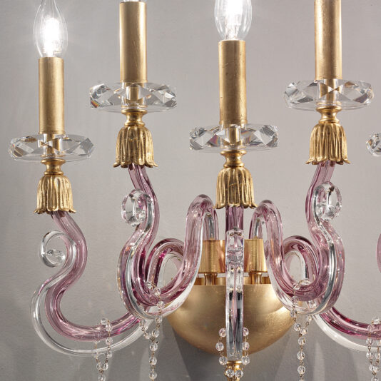 A close-up of the Contemporary 5 Arm Candle Style Glass Wall Light showcases its elegant gold accents and curved, translucent pink glass arms. The chandelier is adorned with crystal ornamentation and lighted bulbs on top, casting a warm glow against a neutral grey wall.