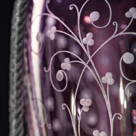 A close-up of the Contemporary Candle Style Glass Chandelier reveals a purple glass fixture adorned with intricate white floral and vine patterns etched into its surface. The chandelier's glossy finish reflects light, accentuating the delicate design details characteristic of contemporary decor. A braided, rope-like texture is visible along the edges, adding to its sophisticated charm.