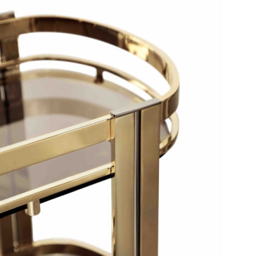 Close-up of the Contemporary Curved Drinks Trolley, featuring a sleek gold-colored metal frame with smooth edges. The modern design includes a top shelf and a glimpse of the partial lower shelf. Nearby, a contemporary velvet bar stool complements the chic aesthetic of the trolley. The glass panels provide a clear, light-reflective surface.
