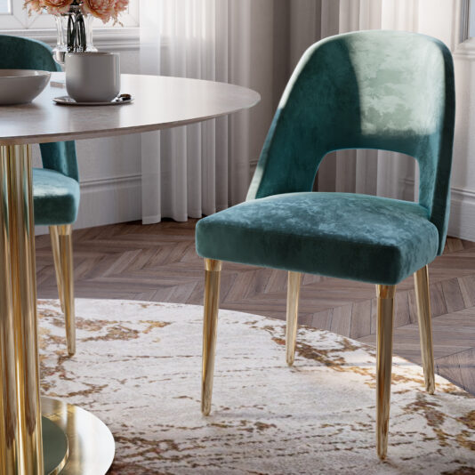A stylish dining room features an elegant round table with a white surface and golden base. In front of the table, a Contemporary Teal Dining Chair with gold legs sits on a light-patterned area rug over herringbone wood flooring. Sheer curtains hang in the background.
