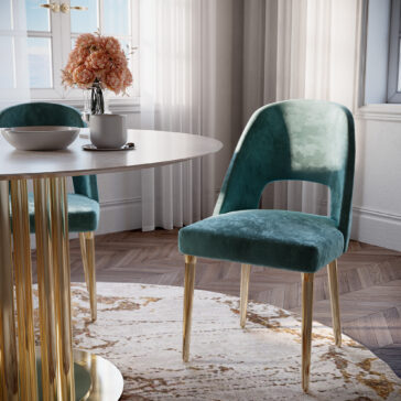 A contemporary dining area showcases a round table with a golden pedestal base, two Contemporary Teal Dining Chairs with golden legs, a white mug, and a white bowl. A clear vase with pink flowers sits in the center. The room boasts large windows, light-colored walls, and a patterned rug.
