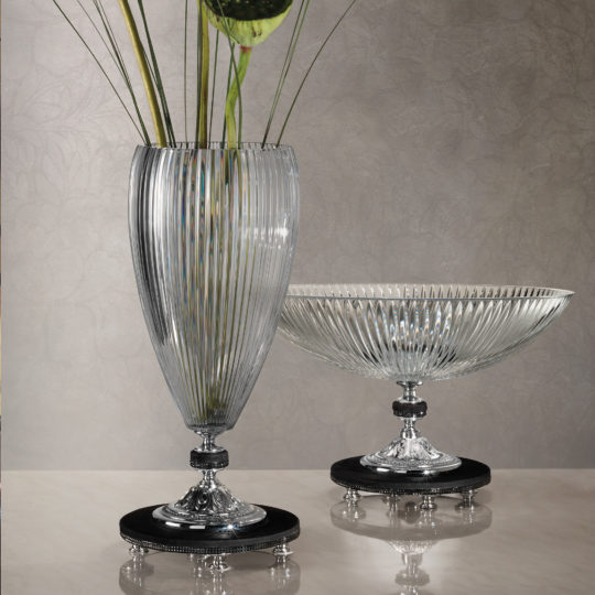The Cut Crystal Centrepiece and Vase, featuring a tall ribbed glass vase with long green stems, stand elegantly on ornate decorative silver bases. They are set against a subtly patterned, neutral-toned background on a reflective surface.