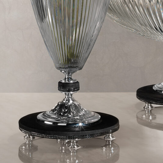 Cut Crystal Centrepiece and Vase A close-up of two intricately designed Cut Crystal Centrepiece and Vases on an elegant black and silver stand. The vases feature vertical grooves, a textured pattern, and ornate silver detailing on the base. The background is neutral with a subtle light pattern.