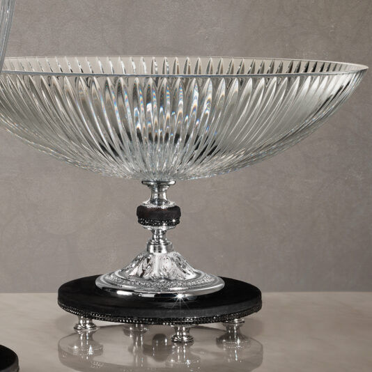 Cut Crystal Centrepiece and Vase The Cut Crystal Centrepiece and Vase, featuring a ribbed design, is elegantly showcased on an ornate and shiny silver pedestal adorned with intricate details. It stands on a polished dark circular base, set against a plain, neutral backdrop.