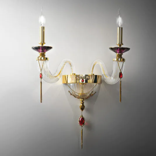 The Double Candle Style Wall Light With Swarovski® Crystals offers a luxurious and ornate design, featuring two candle-shaped bulbs. This elegant sconce is adorned with gold finishes, decorative red gemstones, and gracefully curved glass arms, all mounted on a white wall. Its vintage-inspired aesthetic exudes opulence and sophistication.