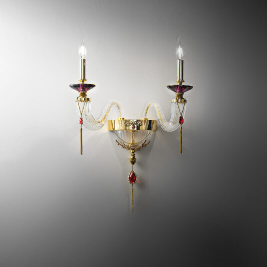 Double Candle Style Wall Light With Swarovski® Crystals The Double Candle Style Wall Light With Swarovski® Crystals features two lit bulbs, a gold base, and intricate design elements. It boasts curved, translucent arms adorned with red accents and is mounted against a plain, light gray wall. Emitting a warm, soft glow, the sconce enhances its elegant appearance.