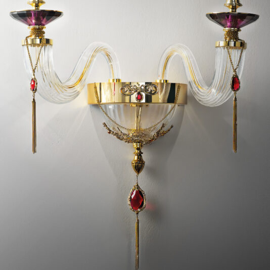 Double Candle Style Wall Light With Swarovski® Crystals The Double Candle Style Wall Light With Swarovski® Crystals is an opulent fixture boasting gold accents and clear, wave-like glass arms. This luxurious sconce features two candle-style lights adorned with red crystal embellishments and dangling gold chains, creating an elegant design mounted on a smooth, light-colored wall.