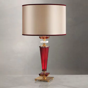 Elegant Ruby Glass Table Lamp The Elegant Ruby Glass Table Lamp features a golden base, a red glass stem, and a beige cylindrical lampshade with red trim accents, all gracefully positioned on a polished gray surface against a neutral background.