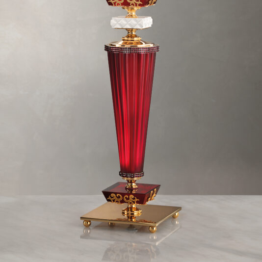 The Elegant Ruby Glass Table Lamp is a luxurious, sophisticated piece featuring a tall, conical red body accented with gold and crystal details. It stands on a square base with ornate golden feet, all set against a neutral background.