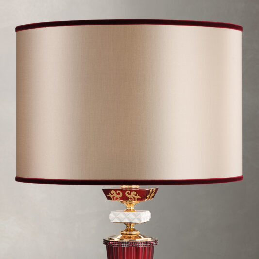 The Elegant Ruby Glass Table Lamp features a wide, cylindrical beige lampshade with red trim at the top and bottom. The ornate base combines gold, white, and red accents with intricate detailing.