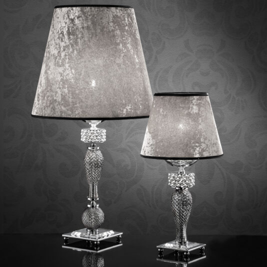Two Glamorous Swarovski® Crystal And Pearl Table Lamps with metallic bases and textured, silver-gray lampshades. The larger lamp stands to the left with a taller, slender stem, while the smaller lamp mirrors the design on a reduced scale. They are displayed against a dark, patterned backdrop.