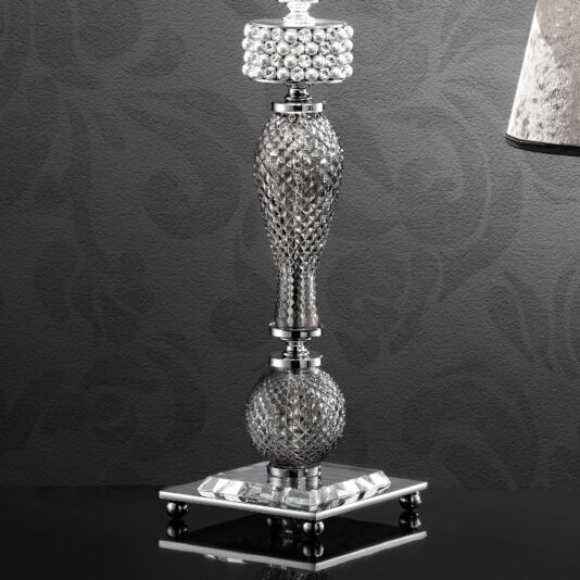 The Glamorous Swarovski® Crystal And Pearl Table Lamp is elegantly positioned on a black surface, set against a dark, patterned wall. Its finely crafted base features textured, diamond-like shapes that culminate in a cylindrical top adorned with crystal-like decorations. A portion of the lampshade is visible on the right.