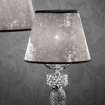 A close-up of the Glamorous Swarovski® Crystal And Pearl Table Lamp, showcasing its textured gray lampshade and an ornate base adorned with Swarovski® crystals. The background features a gray, patterned wall, and a partial view of another similar lamp is visible in the upper left corner.