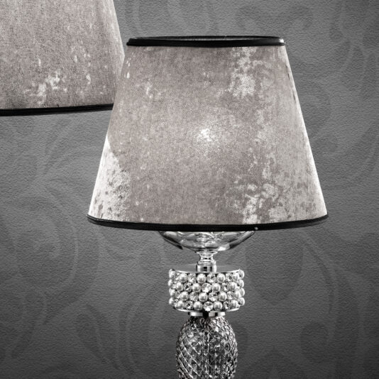 A close-up of the Glamorous Swarovski® Crystal And Pearl Table Lamp, showcasing its textured gray lampshade and an ornate base adorned with Swarovski® crystals. The background features a gray, patterned wall, and a partial view of another similar lamp is visible in the upper left corner.
