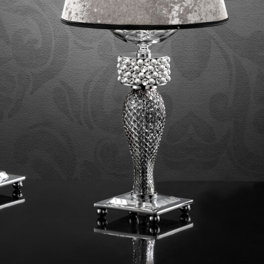 The Glamorous Swarovski® Crystal And Pearl Table Lamp features an intricate silver base with a diamond-patterned texture and a spherical, pearl-like embellishment near the top, standing on a reflective black surface. Accented with Swarovski® crystals, it boasts a grey, velvety lampshade set against a dark, patterned background.