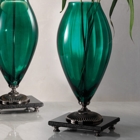 Green Glass Table Lamp And Vase With Rhinestones A decorative scene featuring two large, emerald green glass vases on black bases with metallic accents and detailed designs. A single green leaf extends out of one vase, adding a touch of natural elegance to the sophisticated display, reminiscent of the Green Glass Table Lamp And Vase With Rhinestones.
