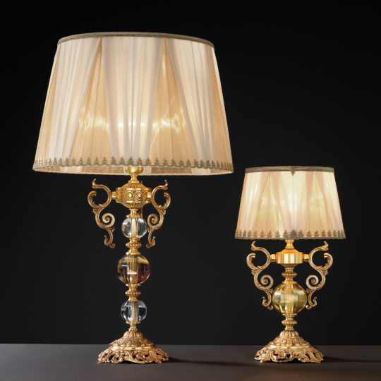A pair of High End Ornate Glass Table Lamps stand elegantly against a dark background. Each lamp features an elaborately designed golden base adorned with intricate scrollwork and clear, spherical ornaments, all topped with pleated beige lampshades that emit a soft, warm glow.