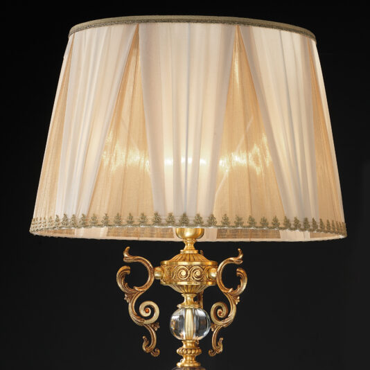 High End Ornate Glass Table Lamp A detailed view of the High End Ornate Glass Table Lamp, featuring a translucent, cream-colored lampshade adorned with gold detailing. The base is elegantly crafted with intricate scrolls and a crystal accent, standing out against a black background.