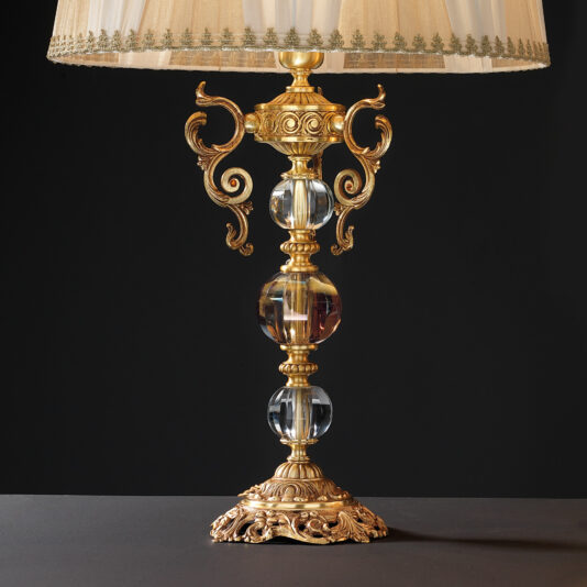 High End Ornate Glass Table Lamp The High End Ornate Glass Table Lamp features a vintage-style design with an ornate gold base and intricately curved decorations. Two large embedded glass spheres accentuate the stem, while a pleated fabric shade with gold trim adds a touch of elegance, all set against a dark background.