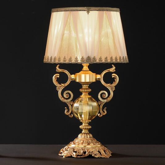 High End Ornate Glass Table Lamp The High End Ornate Glass Table Lamp boasts intricate scrollwork and a round, faceted glass centerpiece. This luxurious item includes a beige lampshade with gold trim, casting a warm, soft glow. The lamp is supported by a richly detailed base and set against a black background.
