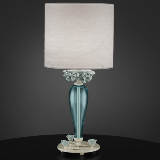 The High End Porcelain Rose Table Lamp boasts an elegant design with a large white cylindrical lampshade, a translucent blue glass base, and decorative white ceramic roses embellishing the neck and base, all set against a dark background.