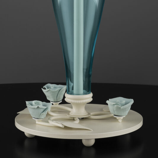 High End Porcelain Rose Table Lamp A sophisticated High End Porcelain Rose Table Lamp features a blue glass vase containing a single white candle, perched on an intricate porcelain base. The base is decorated with delicate pale blue ceramic roses and white leaf accents, creating an elegant display. This exquisite piece sits on a black surface against a dark background, epitomizing elegance and charm.