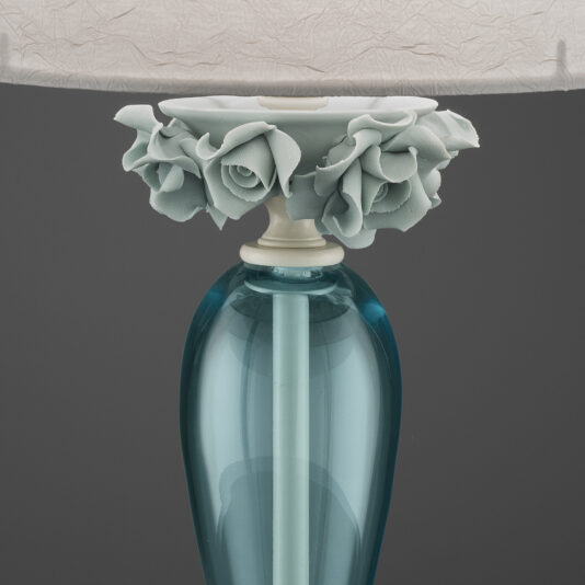 High End Porcelain Rose Table Lamp Close-up of the upper part of the High End Porcelain Rose Table Lamp. The lamp features a white, textured lampshade and a base crafted from blue glass. Where the shade meets the base, intricate sculpted rose flowers in light green add an elegant touch.