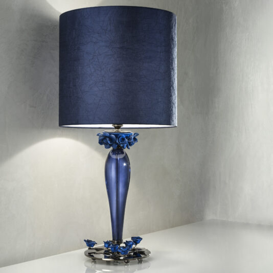 The elegant High End Table Lamp With Porcelain Flowers showcases a stylish blue cylindrical lampshade and a sleek glass base adorned with intricate decorative porcelain flowers. Placed on a white surface against a light gray wall, the lamp highlights its exquisite design and craftsmanship.