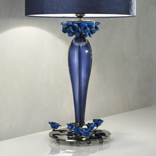 The High End Table Lamp With Porcelain Flowers features a sleek, elongated blue glass base adorned with intricate porcelain flowers near the top and bottom, complemented by a large blue lampshade. It stands on a polished surface against a smooth, light-colored wall.