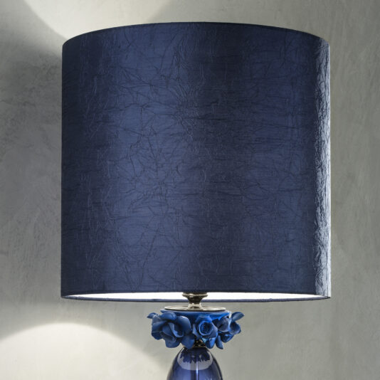 A close-up of the High End Table Lamp With Porcelain Flowers showcases its dark blue, textured lampshade. The base, adorned with a cluster of blue porcelain flowers, adds an elegant touch. The lamp is lit, casting a soft glow on the wall behind it.