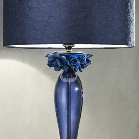 A High End Table Lamp With Porcelain Flowers, boasting a deep blue base and a textured navy blue lampshade. The base is adorned with intricate porcelain flowers around the metal fixture that connects the shade to the base. This elegant lamp stands against a smooth beige wall.