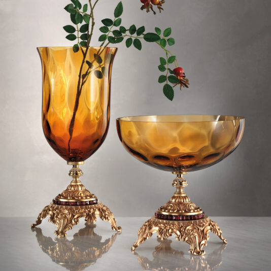 The Jewelled Amber Glass Centrepiece and Vase feature a tall vase and matching bowl on ornate, gold-colored stands. The vase holds a green leafy branch adorned with two small ruby-colored fruit ornaments. Both pieces rest on a white marble surface against a grey background.