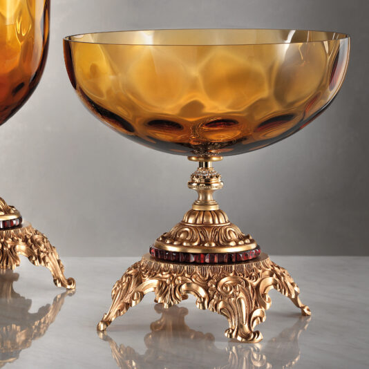 A close-up view of the Jewelled Amber Glass Centrepiece and Vase showcases two ornate amber glass bowls with gold-accented bases. The intricate designs on the bases, featuring swirling patterns and red accents, lend a luxurious and antique appearance to these elegant bowls. The background is a soft, neutral gradient, enhancing the charm of this jewelled masterpiece.