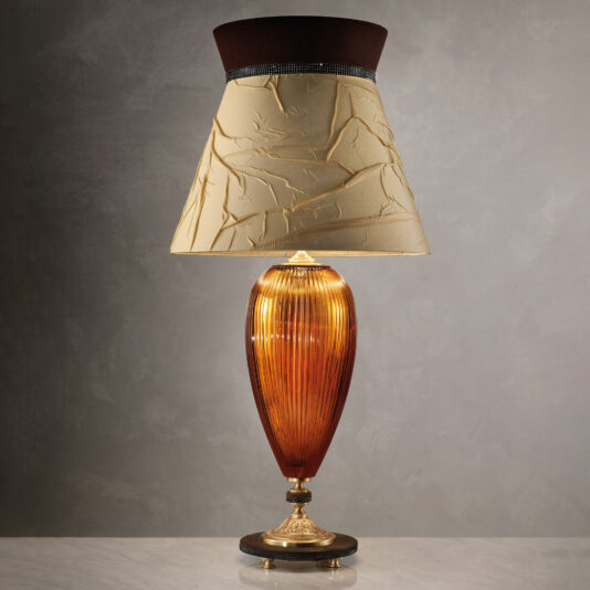 The Large Contemporary Amber Cut Crystal Table Lamp boasts a textured beige lampshade with a dark brown top edge. Its sleek, oval-shaped amber cut crystal base features exquisite ridged detailing and stands gracefully on an ornate brass pedestal, set against a neutral background.