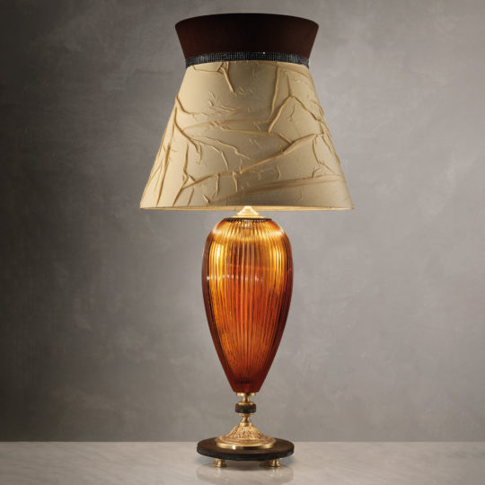 The Large Contemporary Amber Cut Crystal Table Lamp boasts a textured beige lampshade with a dark brown top edge. Its sleek, oval-shaped amber cut crystal base features exquisite ridged detailing and stands gracefully on an ornate brass pedestal, set against a neutral background.