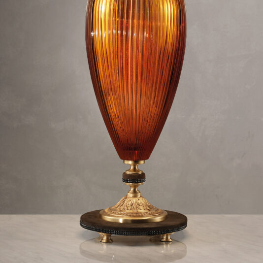 Large Contemporary Amber Cut Crystal Table Lamp The Large Contemporary Amber Cut Crystal Table Lamp, featuring an elegant ribbed glass shade and a brass base with intricate detailing, stands on a marble surface. The backdrop is a neutral grey wall, and the lamp emits a warm glow.