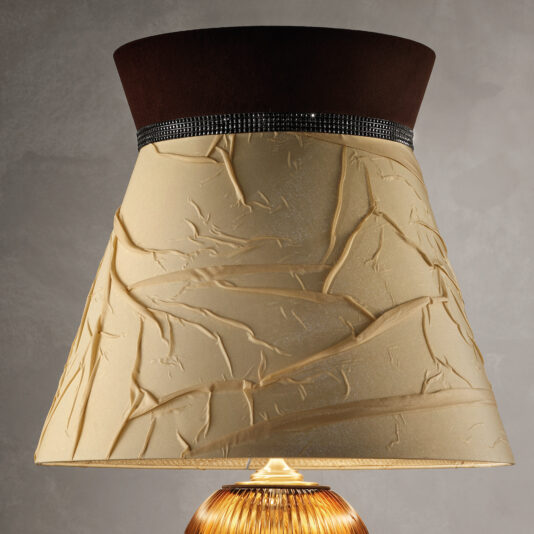 Large Contemporary Amber Cut Crystal Table Lamp A close-up of the Large Contemporary Amber Cut Crystal Table Lamp featuring a unique, cream-colored shade with artistic creases and a dark brown band at the top, adorned with small, sparkling embellishments. The base of the amber cut crystal lamp is partially visible.