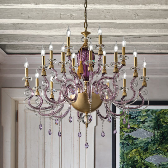 The Large Contemporary Candle Style Glass Chandelier, adorned with gold and purple accents, hangs prominently from the wooden ceiling. It features multiple candle-shaped bulbs and intricate glass arms with teardrop-shaped pendants. The room is decorated with white walls and a framed artwork on one side.