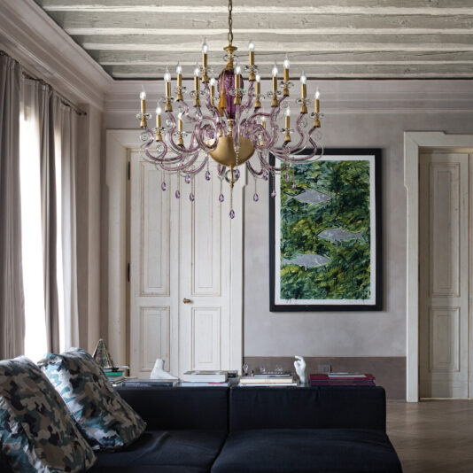 A cozy living room features a gray couch adorned with camouflage cushions, a wooden-beamed ceiling, and the striking Large Contemporary Candle Style Glass Chandelier. Artwork depicting fish accents the light gray walls, while curtains frame a large window. Multiple books and trinkets rest on the coffee table.