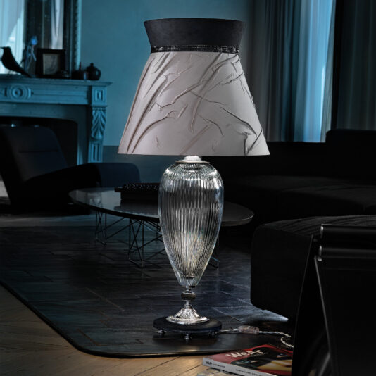 The Large Contemporary Cut Crystal Table Lamp, featuring a fluted glass base and a textured lampshade, is elegantly placed on a dark-toned coffee table in a sophisticated, dimly lit living room. The background highlights modern furniture and ambient natural light streaming through a window.