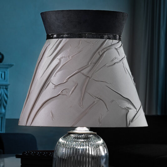 Large Contemporary Cut Crystal Table Lamp Experience elegance with the Large Contemporary Cut Crystal Table Lamp, featuring a textured, artistic lampshade. The lampshade combines light fabric with embossed branch-like patterns and a dark upper band adorned with shiny accents. Its base showcases a metallic, ribbed design against a dimly lit background, reminiscent of contemporary luxury.
