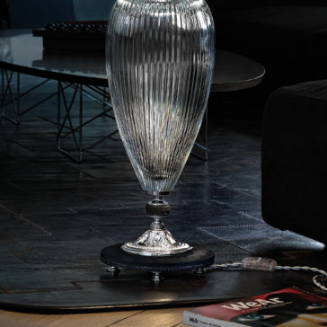 Large Contemporary Cut Crystal Table Lamp A close-up of an elegant, elongated, ribbed glass vase with a silver base. It's placed on a dark wood floor next to a modern black sofa. The Large Contemporary Cut Crystal Table Lamp stands nearby. A magazine with a red cover lies partially on the floor in the foreground. A sleek black coffee table is in the background.