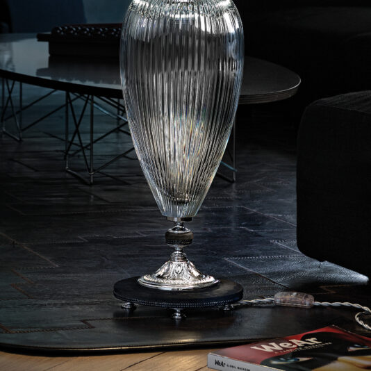 Large Contemporary Cut Crystal Table Lamp A close-up of an elegant, elongated, ribbed glass vase with a silver base. It's placed on a dark wood floor next to a modern black sofa. The Large Contemporary Cut Crystal Table Lamp stands nearby. A magazine with a red cover lies partially on the floor in the foreground. A sleek black coffee table is in the background.