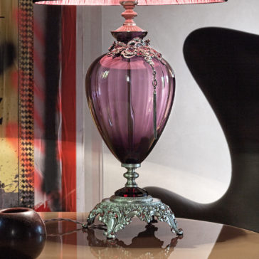 A large ornate jewelled glass table lamp featuring a purple base and an exquisite metal stand is adorned with a pink and white lampshade that complements its elegance. Positioned on a reflective surface, it stands near a dark, abstract-shaped object against a colorful, patterned backdrop.