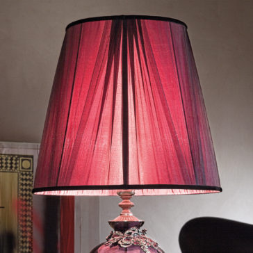 The Large Ornate Jewelled Glass Table Lamp features a stunning, decorative base paired with a pink, intricately detailed pleated lampshade. It creates a soft ambient glow that adds elegance to any room. The jewelled glass on the base enhances its sophisticated look, while the warm reddish light from the pleated lampshade casts an inviting glow against a neutral, plain wall.