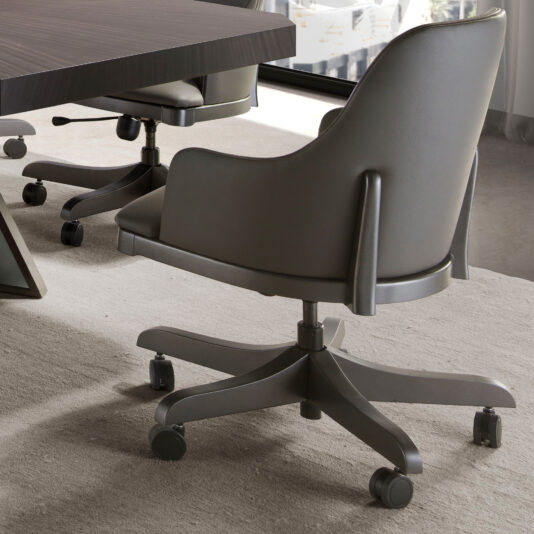 A Leather Swivel Office Chair On Castors with a grey cushioned seat and backrest, attached to a five-wheeled base, is angled near a dark wooden table. The chair sits on a light-colored carpet, with sunlight streaming through the window in the background.